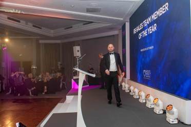 A man in a suit walks on stage at an awards event, with a screen displaying "Sales Team Member of the Year" in the background. Guests are seated at tables in the audience.