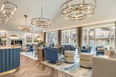 Tennyson's Lounge Bar & Bistro A modern lounge with blue and beige armchairs, round tables, gold light fixtures, large windows, and wood-patterned flooring.