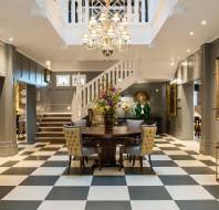 Audley Chalfont Dene Lobby Elegant hotel lobby with black and white checkered floor, central round table with chairs, chandelier, staircase, paintings, and decorative flowers.