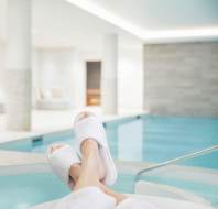 Person wearing a white robe and slippers relaxes by an indoor pool in a bright, modern spa with lounge seating in the background.