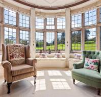 Sunlit sitting area with large bay windows overlooking a green lawn. Two armchairs and a cushioned window seat are arranged on a beige carpet with floral curtains framing the windows.