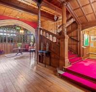 A spacious interior featuring wooden paneling, a central red-carpeted staircase, arched doorway, and polished wood floor. Tables with lamps and a green exit sign are also visible.