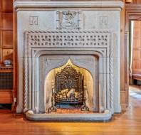 A detailed stone fireplace with an ornate metal grate and wood logs inside, set against a paneled wooden wall in a well-lit room with wooden floors.