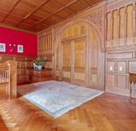A furnished room with wooden paneled walls and ceiling, red wallpaper accent, intricate double door, and parquet flooring. The room includes a decorative rug, furniture, and a staircase with a chandelier.