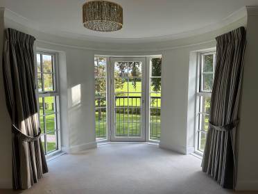 A room with a bay window and gray curtains overlooks a grassy outdoor space. A chandelier hangs from the ceiling.