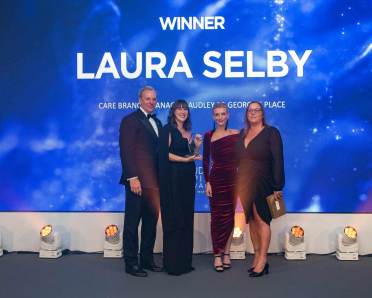 Four people pose on stage with a trophy in front of a large screen displaying "Winner Laura Selby.