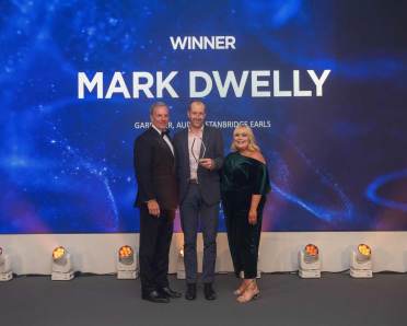 Three people stand on stage with one holding an award. A large display in the background reads "Winner Mark Dwelly.