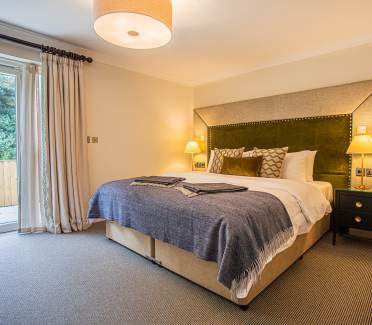 A neatly arranged bedroom with a large bed. There are lamps on nightstands, a large window with curtains, and a gray carpet covers the floor.