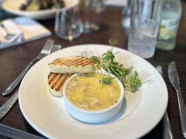 A small dish of pâté with two slices of toasted bread and greens on a white plate, placed on a dark wooden table with glasses and utensils nearby.