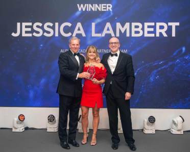 Three people in formal wear on stage hold a trophy. The background displays "Winner Jessica Lambert" with abstract blue graphics.