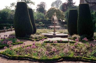 A formal garden with a central fountain, surrounded by shaped hedges and blooming flowers. Paths and neat garden beds are visible with trees in the background.