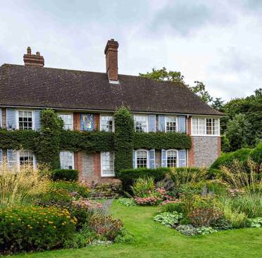 A two-story brick house with ivy-covered walls and blue shutters is surrounded by a lush garden with various plants and flowers.