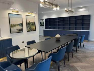 A modern conference room with a long table, blue chairs, a small round table, framed landscape art, a bookshelf, and a chandelier.