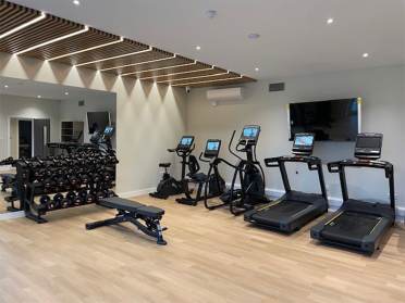 A modern gym with treadmills, stationary bikes, dumbbells, a bench, and a wall-mounted TV.