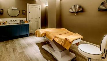 A massage room with a treatment table, covered with a yellow towel, a sink with toiletries, and decorative wall fans.