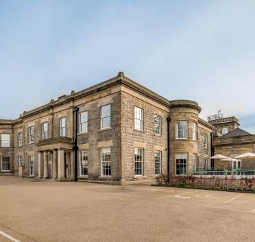 A large, rectangular stone building with numerous windows, columns by the entrance, and a flat roof, set in an open paved area.