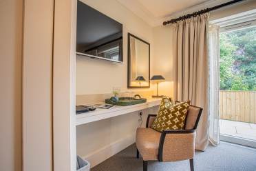 A modern hotel room corner with a wall-mounted TV, a desk, a lamp, and a cushioned chair. A sliding glass door opens to a wooden fenced balcony, and neutral-colored curtains hang beside it.