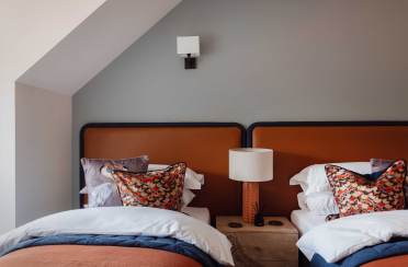 A bedroom with two single beds, each with white and colorful pillows, shares a wooden nightstand with a lamp. The walls are painted gray, and there's a wall light above the beds.