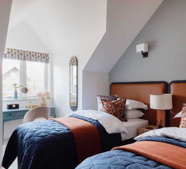 A cozy bedroom features two twin beds with blue quilts and orange throws, a blue desk by the window, and a mirror on the wall. A lamp and decorative pillows complete the space.