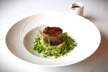 A gourmet dish featuring a cylinder of cooked meat on a bed of peas and greens, with a sprig garnish, served on a white plate. A small sauce pitcher is in the background.