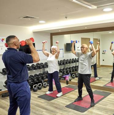 Two people participate in a fitness class, holding dumbbells overhead, guided by an instructor. They are in a gym with mirrored walls and various weights.