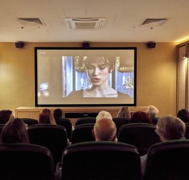 People in a dimly lit room watch a film on a large screen, featuring a woman in period costume.