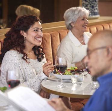 Two women sit at a restaurant table enjoying a meal and conversation. One holds a glass of red wine. A man is in the foreground, partially facing away, looking at a menu.