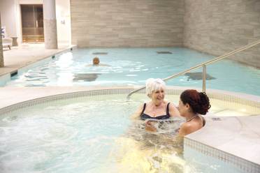 Two people relax in a jacuzzi talking, while another person swims in a larger pool in a spa setting.