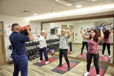 A fitness instructor guides three people lifting dumbbells in a gym mirrored room.