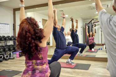 Four people in workout clothes perform yoga poses on mats in a gym with mirrored walls and gym equipment in the background.