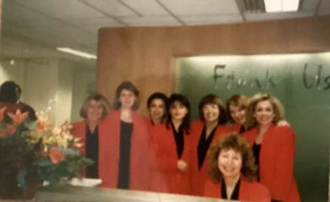 Group of eight women in red blazers, smiling, standing indoors in front of a wall with cursive writing. A floral arrangement is visible in the foreground.