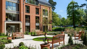 Phase II at Sunningdale Park A modern brick apartment building with balconies overlooks a landscaped courtyard featuring benches, young trees, and various potted plants on a sunny day.