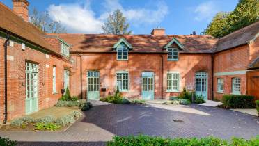 The Stables - Exterior A red-brick house with green doors and windows, surrounded by a landscaped courtyard and greenery.