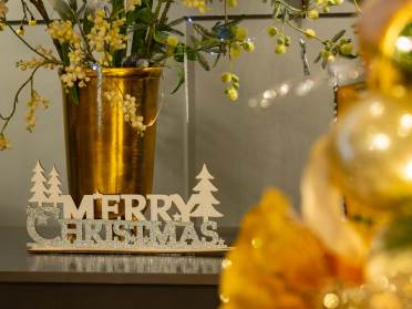 Christmas Fair at an Audley Village A glittery "Merry Christmas" sign sits on a surface next to a gold vase with festive greenery; blurred gold decorations appear in the foreground.
