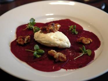 Beetroot carpaccio and goat’s cheese mousse A white plate with beetroot slices, a dollop of cream, candied nuts, and microgreens.