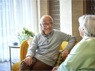 Owners lifestyle  Two elderly people sitting on a yellow sofa, engaged in conversation, with light shining through sheer curtains in the background.