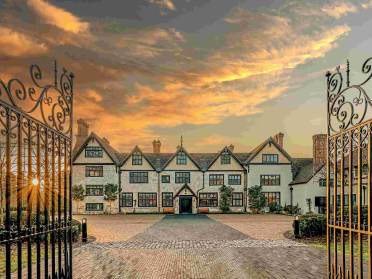 Golden hour at Audley Stanbridge Earls A large Tudor-style mansion with multiple chimneys is seen through open wrought iron gates at sunset, with a cobblestone driveway leading to the entrance.