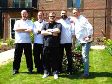 Audley restaurants Chefs Five men in chef uniforms stand together on a sunny lawn in front of a brick building, posing and smiling at the camera.