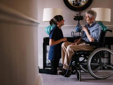 Laura Selby A nurse kneels beside an elderly man in a wheelchair, smiling and engaging in conversation in a well-lit room with lamps and a mirror.