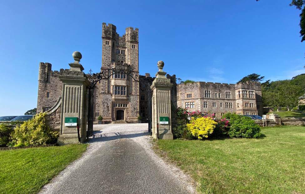 A historic stone castle with two tall towers, an arched entrance, and a gravel driveway, surrounded by manicured gardens and lawns under a clear blue sky.