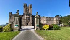 A historic stone castle with two tall towers, an arched entrance, and a gravel driveway, surrounded by manicured gardens and lawns under a clear blue sky.